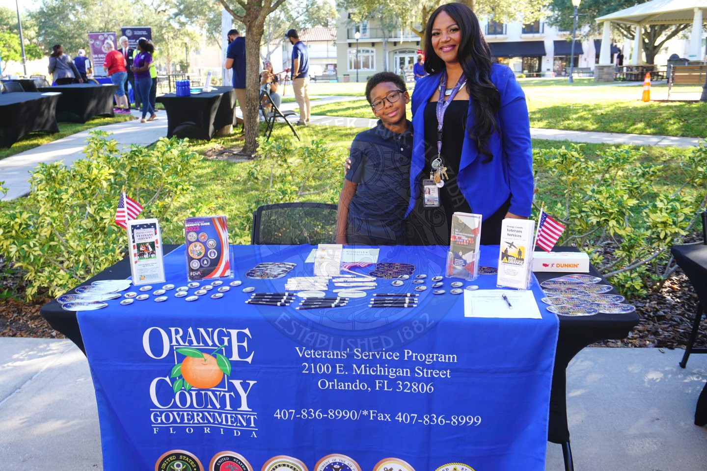 A table that has a blue table cover saying Orange County Government with some informational products laying on top of it. There is a lady with a boy  wrapped around her arm smiling.