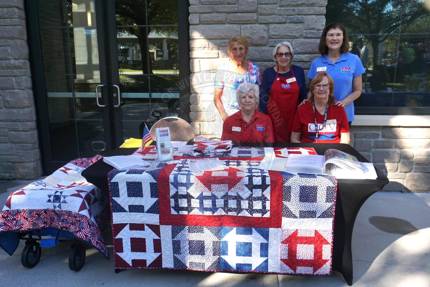 a table with an american themed quilt laying on tob of the black linen table cloth with a group of 5 women stnading behind the table smiling.