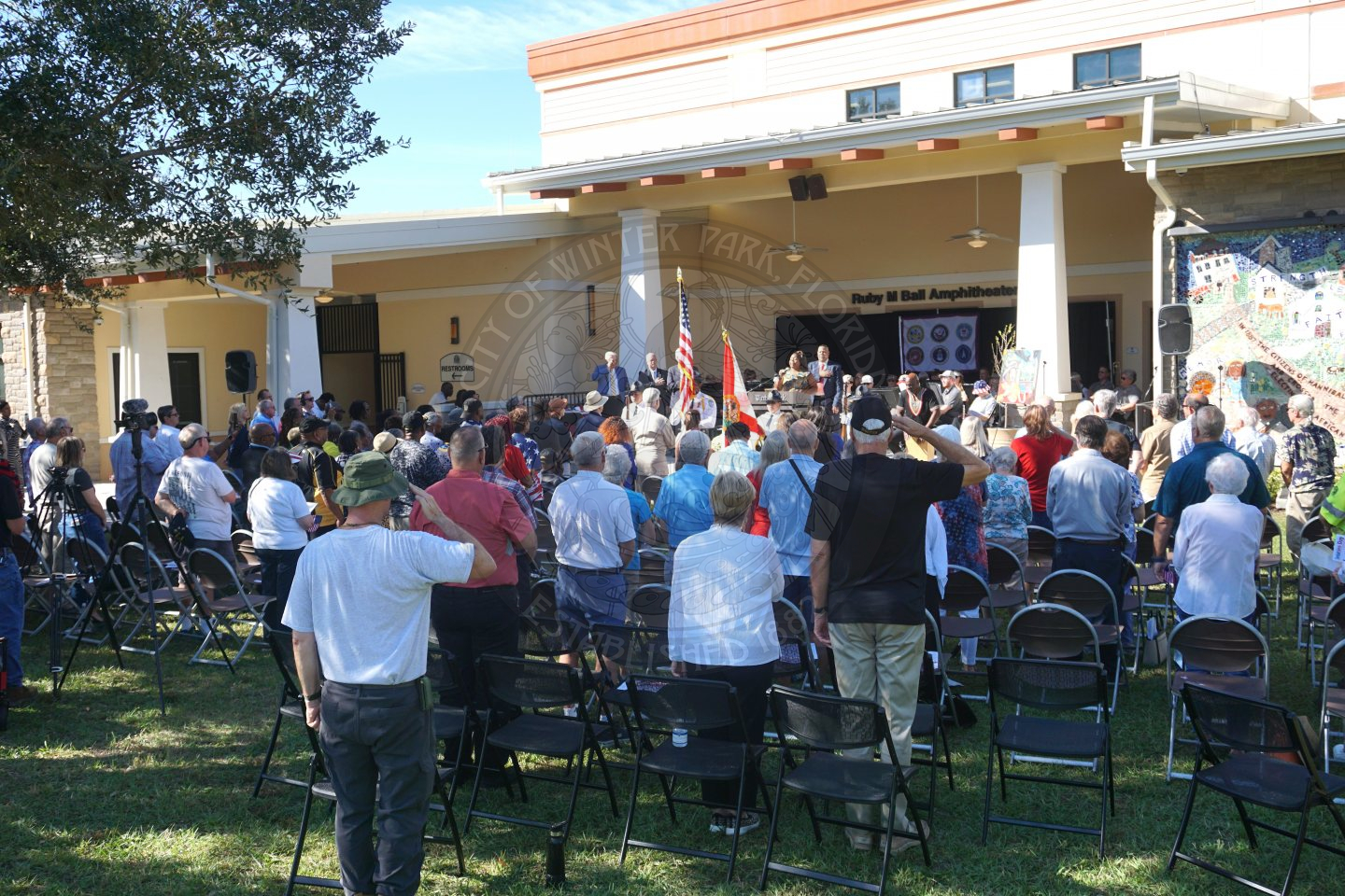 People standing in front of their chairs saluting to the Winter Park Police Department Color Guard.