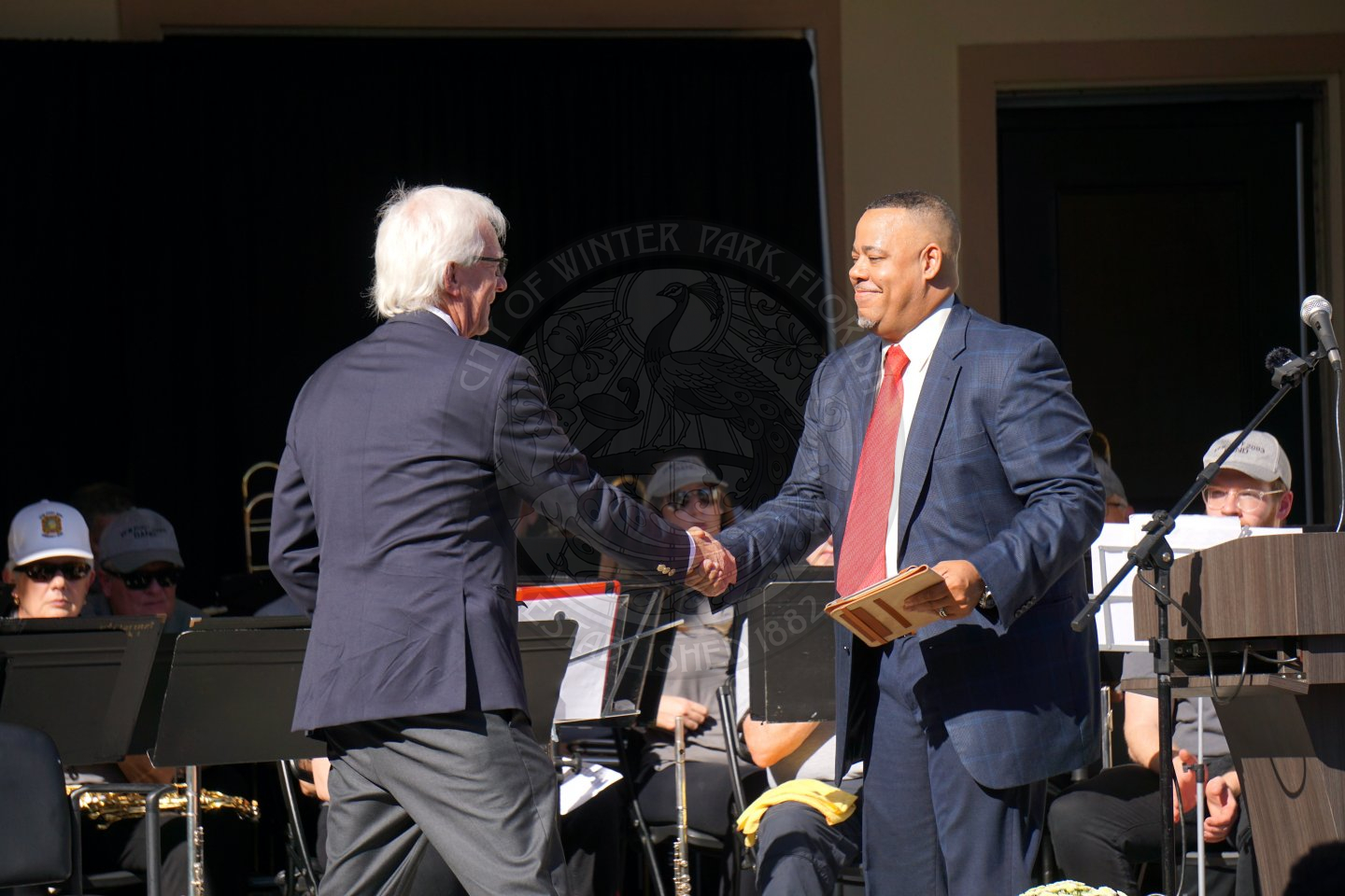 2 men wearing business suits shaking hands while standing on a stage with people sitting behind them playing instruments.