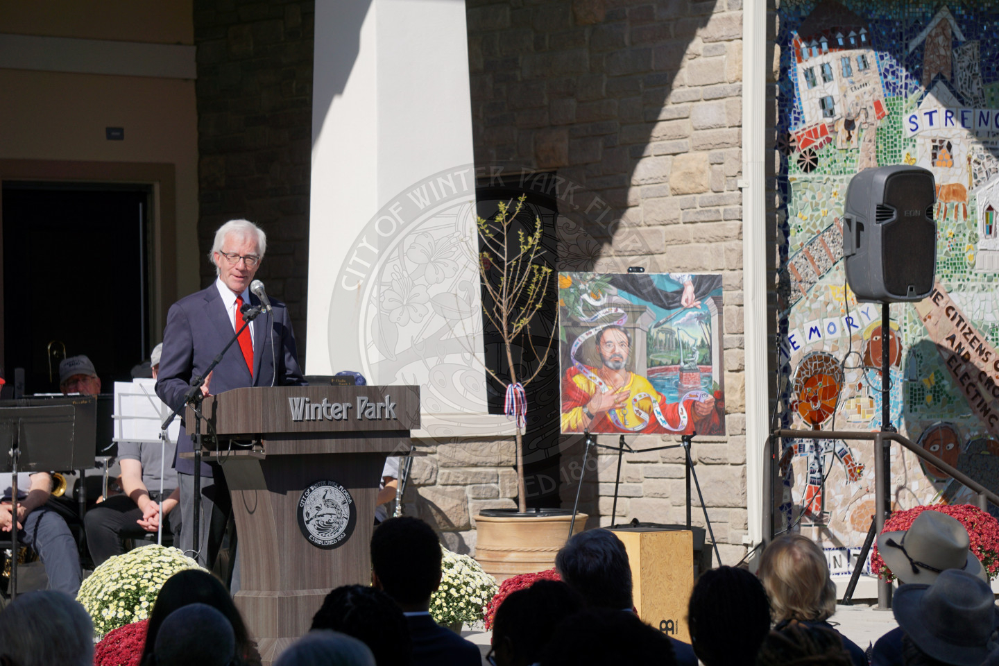 The Mayor of Winter Park standing on stage behind a podium that says Winter Park speaking into a mircophone in front of people. To the right of the mayor is a picture that is on an easel with a tree next to it.