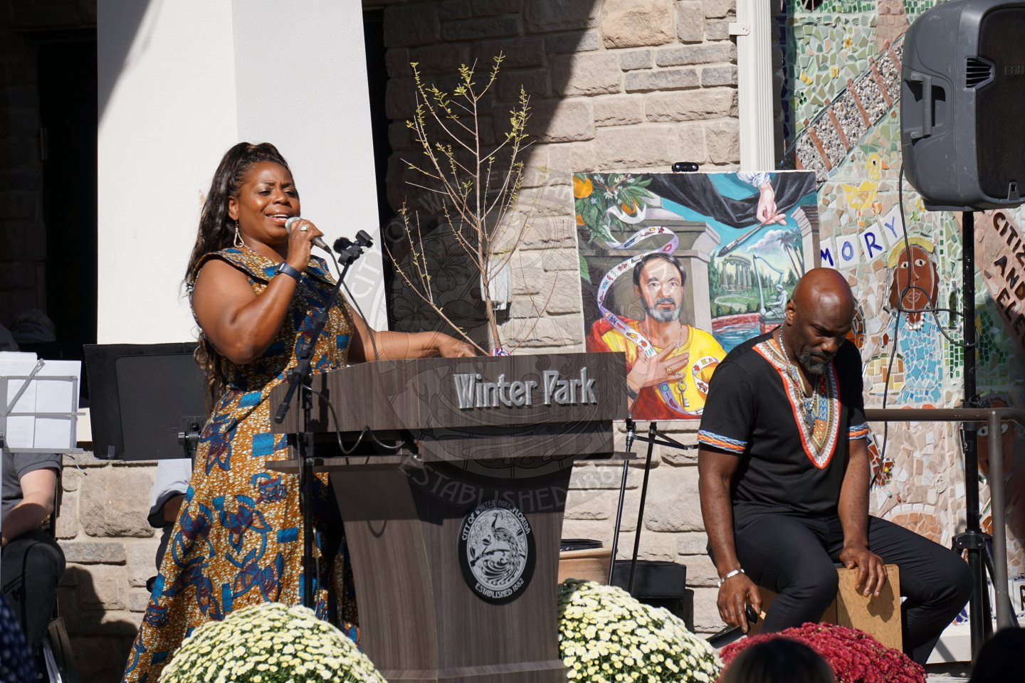 Woman standing behind a podium that says Winter Park with the Winter Park city seal underneath singing into a microphone. To the right of the woman is a man standing down playing the drums with his hand on a wooden box.