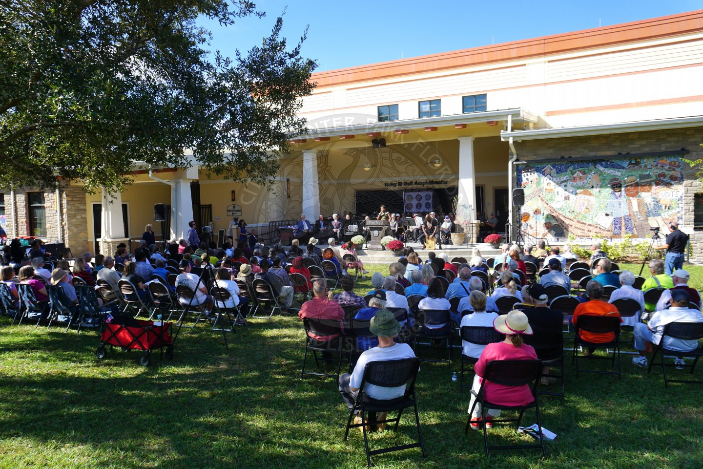 People sitting in rows of chair that are placed in a grass. They are facing. astage with a band playing instruments. On the stage in front of the band is a podium that says Winter Park with a city seal underneath.