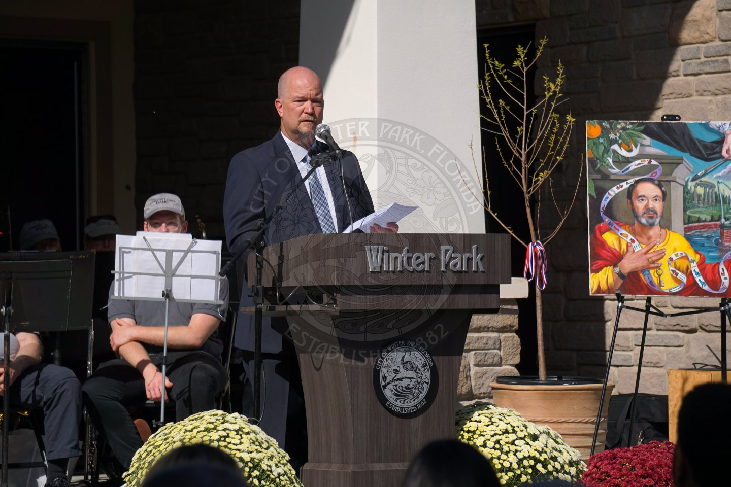 On a stage, there is a man in a business suit standing behind a podium that says Winter Park with the city seal underneath it. There is a painting and a baby tree to the right of the man on the stage.