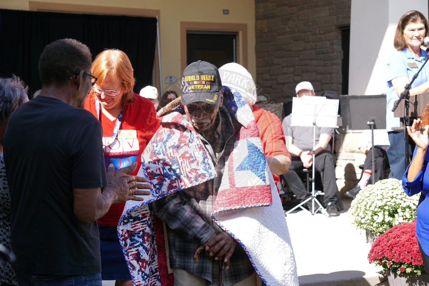 two women placing an american red, white and blue patterend quilt on a mans shoulder with another family member standing on the left side of him.