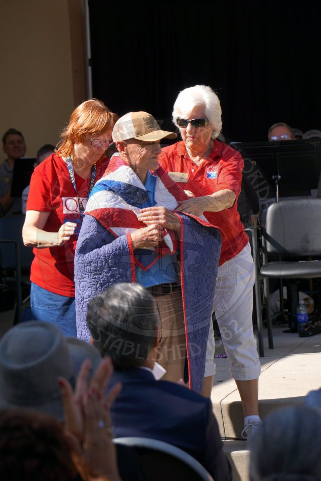 2 women wrapping an american red, white and blue patterend quilt on the shoulders of a man standing in front of them.