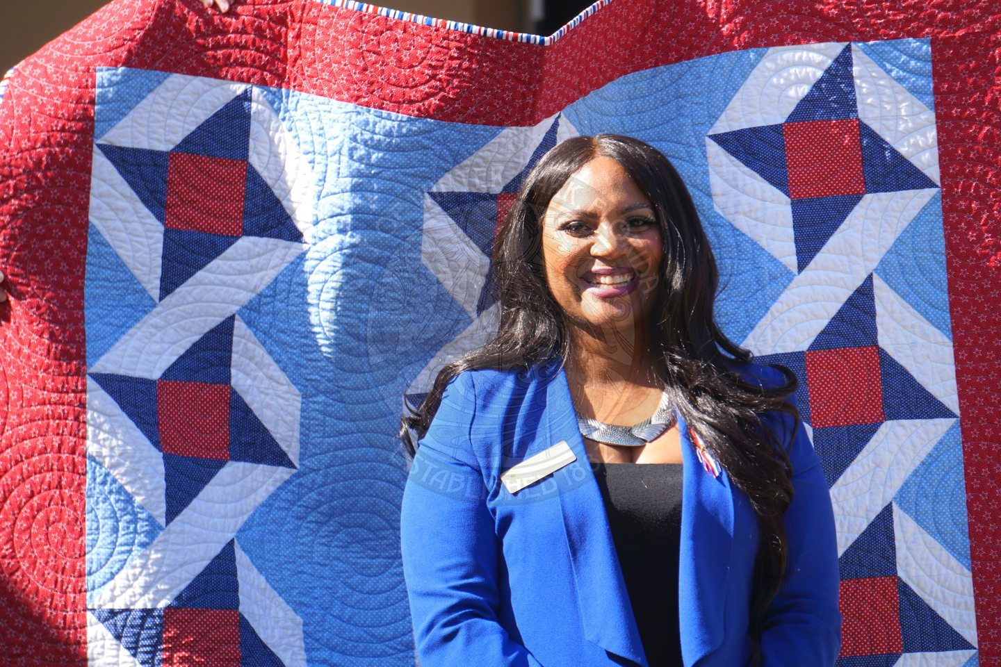 a woman smiling standing in front of an american (red, white and blue) quilt.