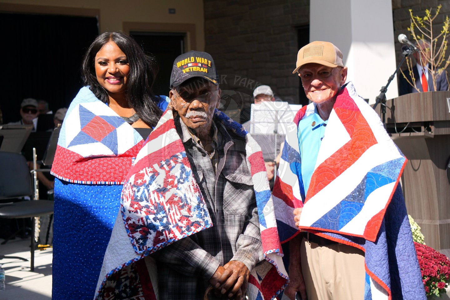 1 man and another man and woman standing behind the man in the middle. they all have american quilts laying on their shoulders that are red, white and blue.