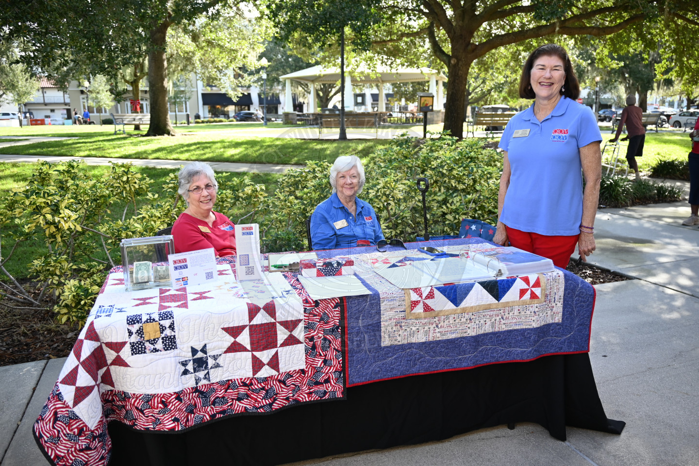 a table filled with two homemade quilts. There are two ladies sitting in chairs behind the table and one lady standing to the side of the table. 