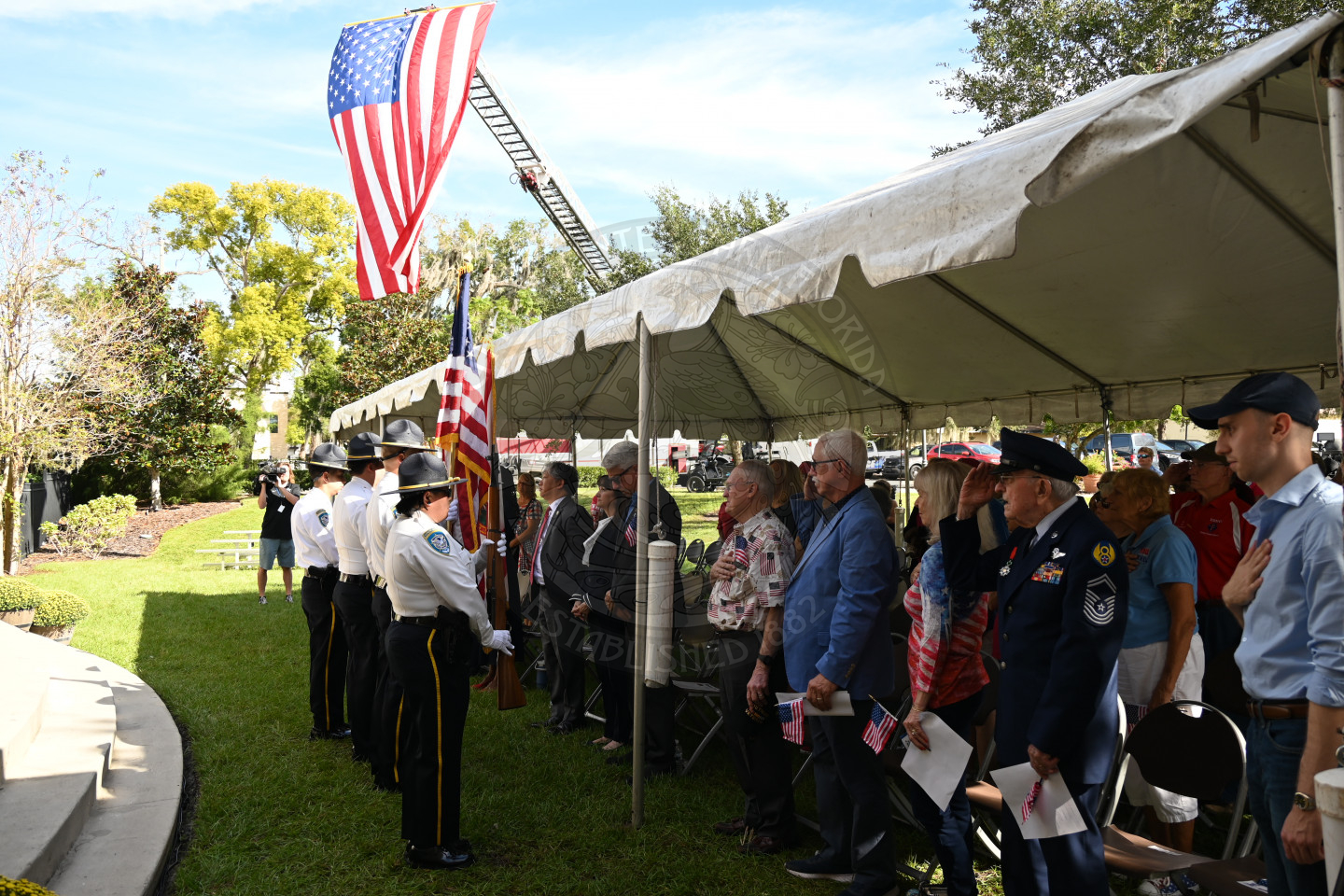the honor guard saluting with their flags right in front of a tent filled with a bunch of people standing up. There is a flag in the background hanging from firetruck. 