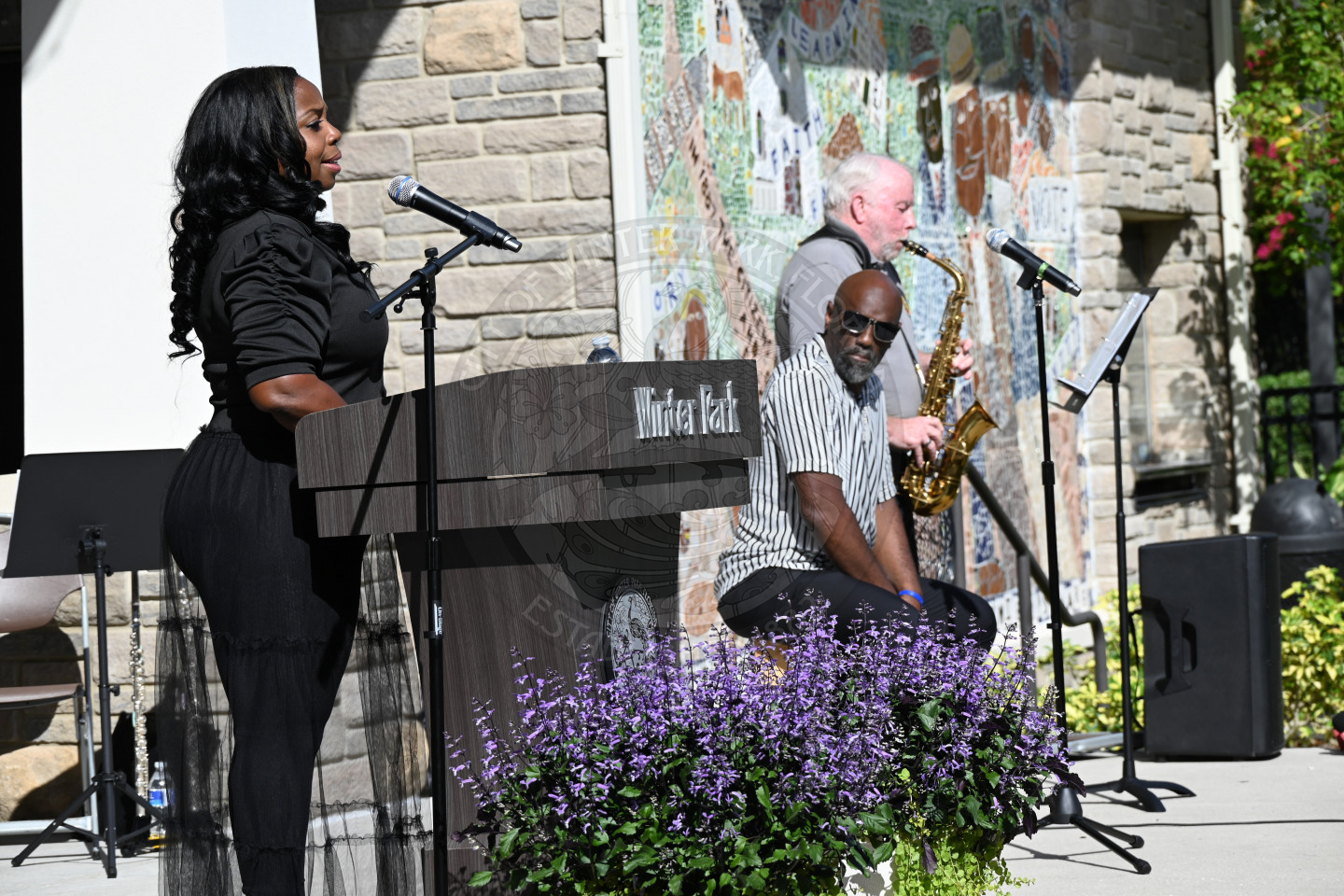 A woman singing into microphone with two man standing to the left of her. One man is sitting down drumming and the other is standing up playing his saxophone. 