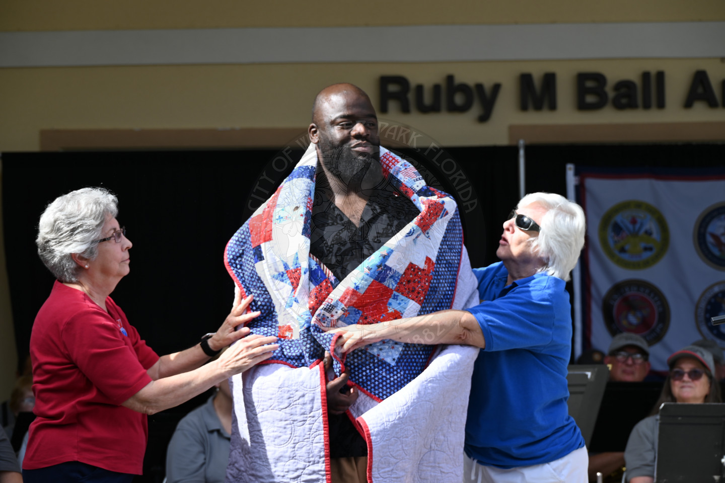 A man standing on a stage accepting his quilt of valor being wrapped around his body.
