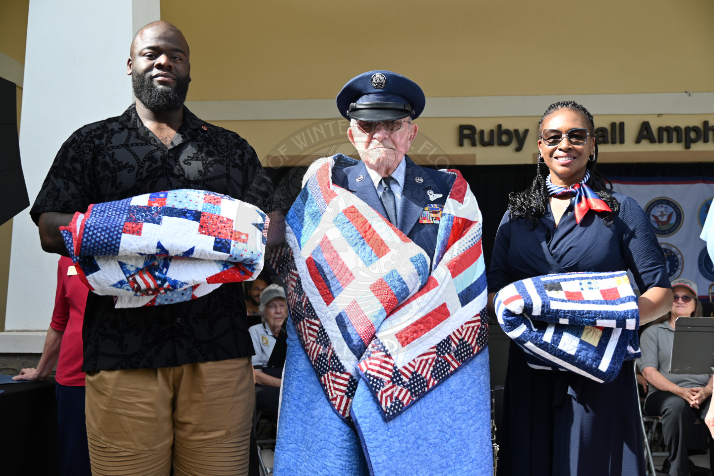 three quilts of valor recipients standing with their quilts on the stage.