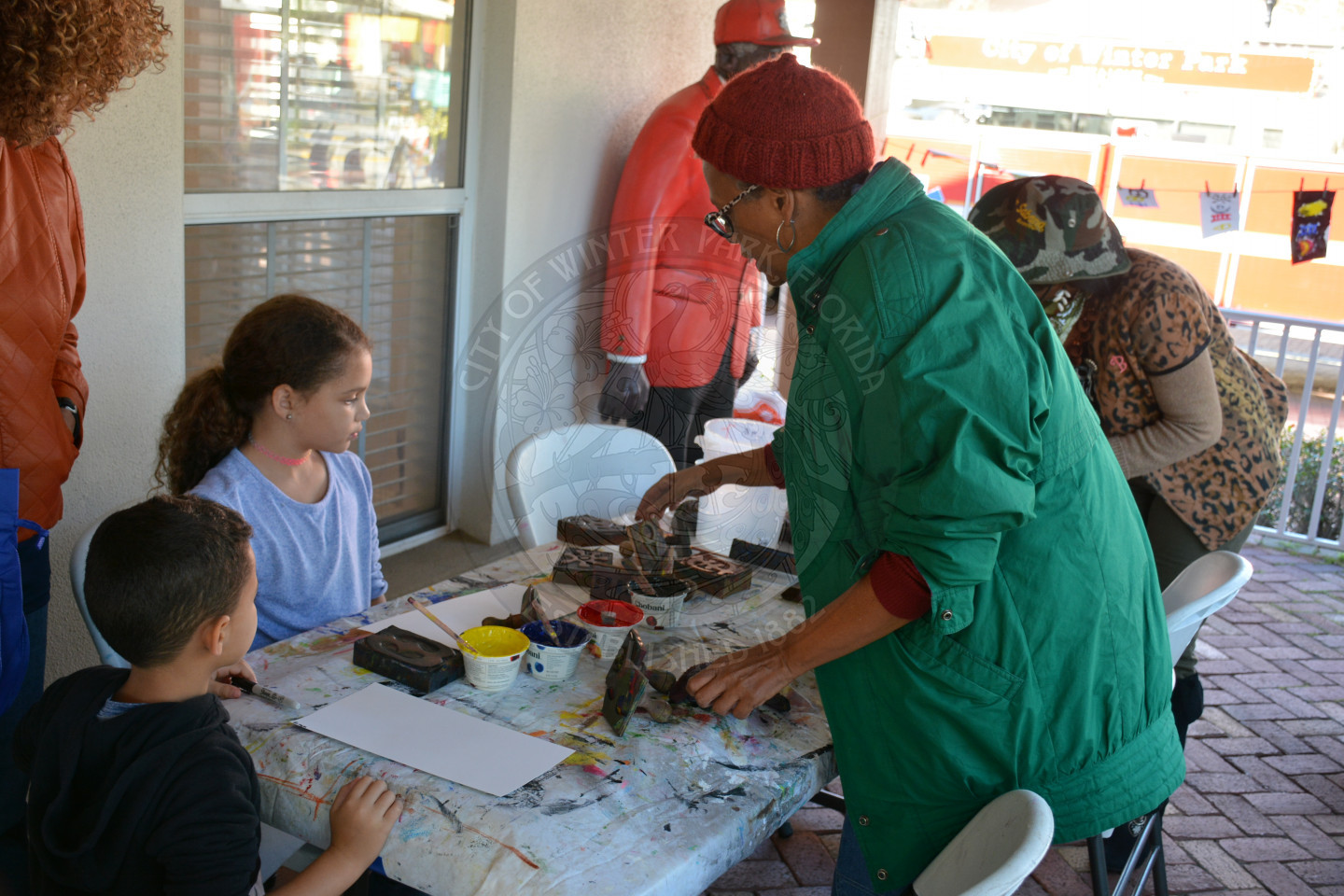 two young children seated at arts and crafts table while female adult helps them with the project