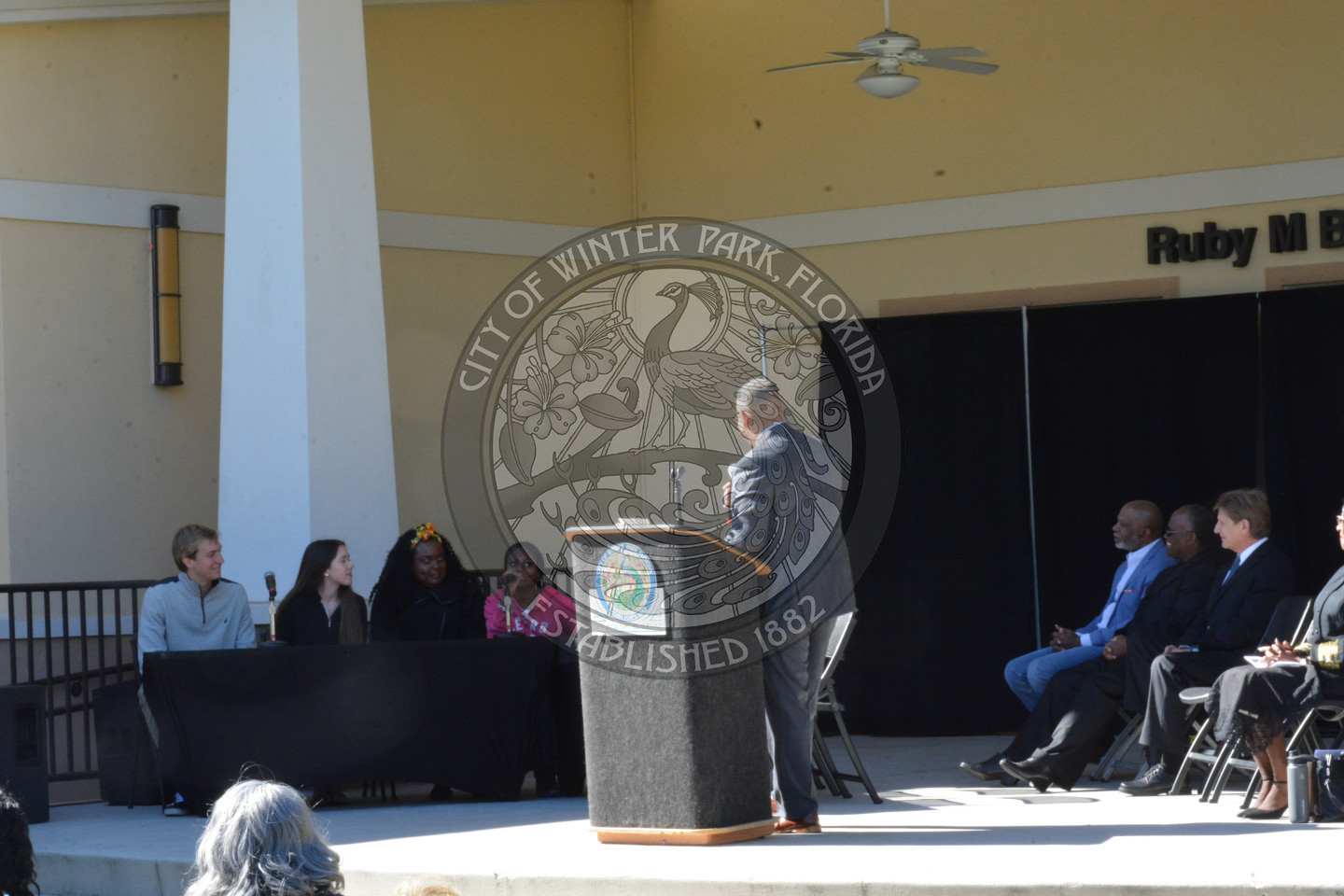 man on stage behind a podium talking to a panel of teens