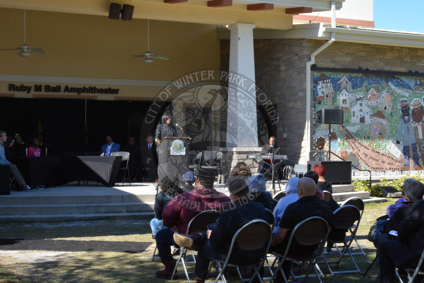 woman on stage preaching behind a podium and a crowd sitting in chair in front of her