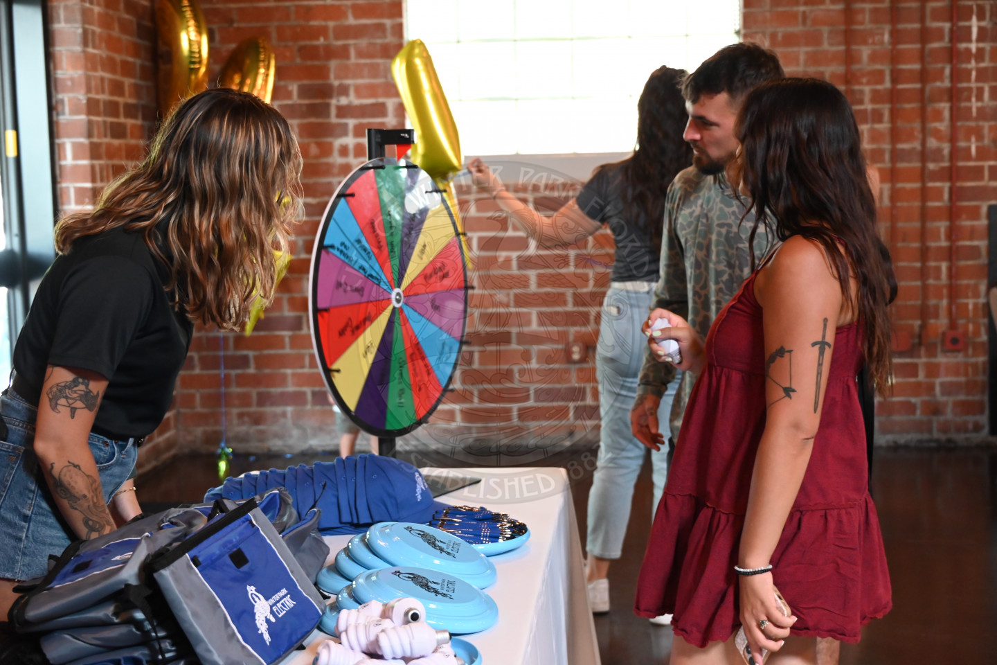 two people spinning the wheel on a table with a lady standing behind the wheel looking over to see where the wheel lands.