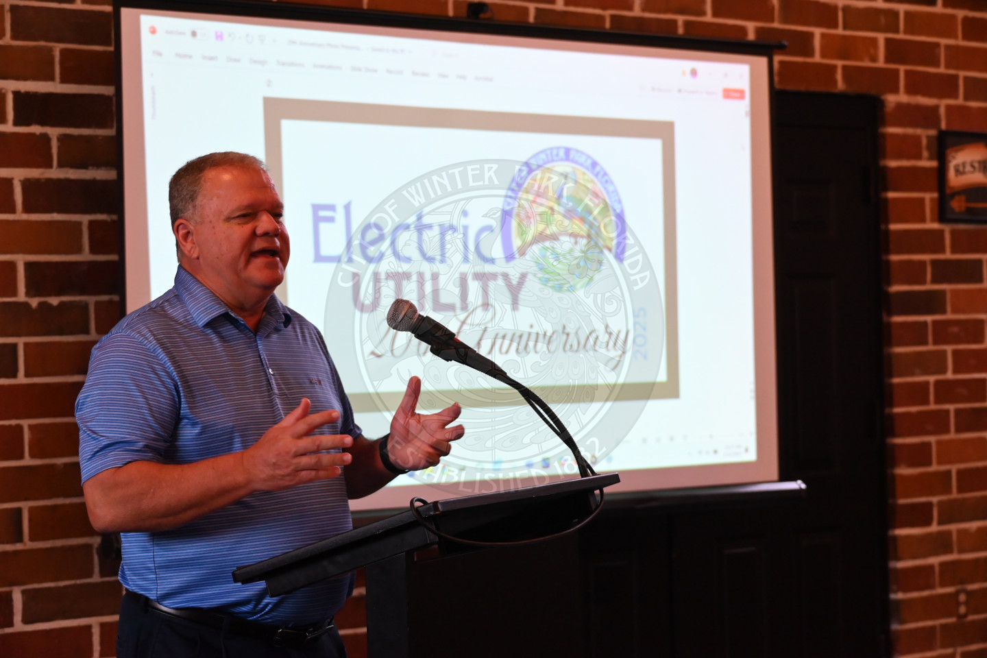 a man speaking into a microphone with a video monitor behind him.