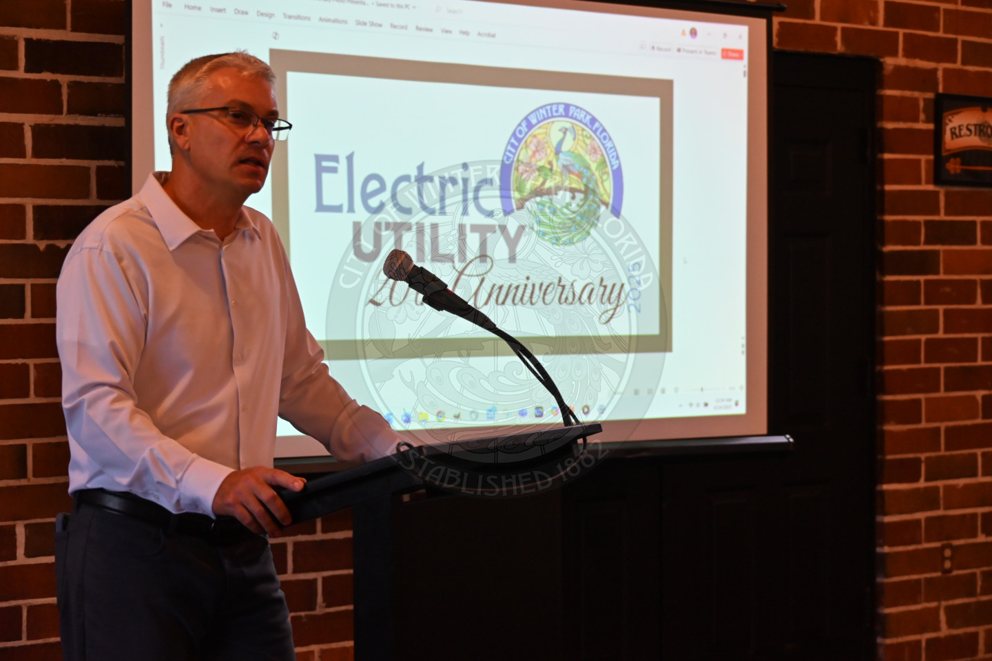 a man speaking into a microphone with a video monitor behind him.