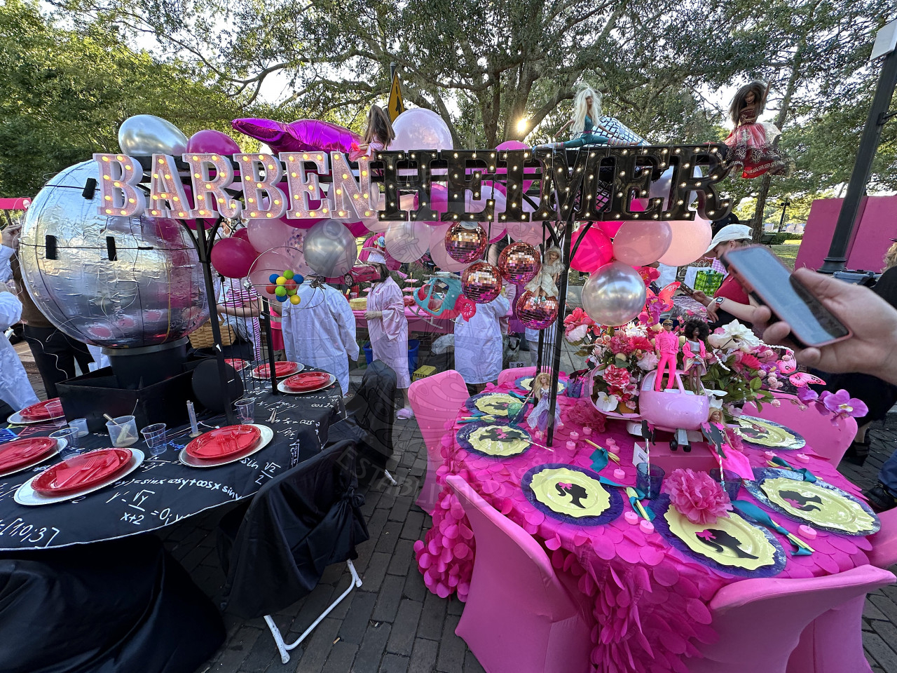 two round tables pushed together to make Barbenheimer which is the movies Barbie and Oppenhiemer put together. One table has scientific equations and a giant metalic globe in the middle and the other table is all pink with barbie dolls and barbie plates on it.