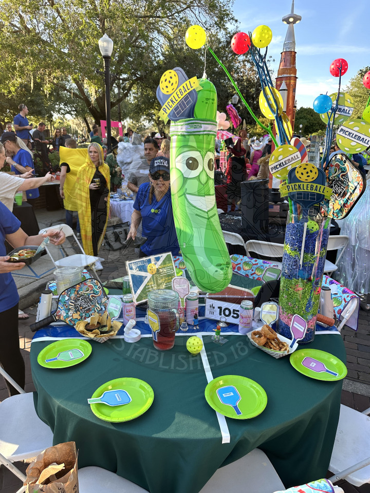 a pickleball themed table. There is a blow up pickle  in the middle of table. around the table and in front of the chairs are paper plate pickleball plates with pickle balls and pickleball paddles on the table.