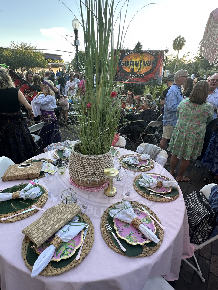 a table with a huge snake looking plant. There is a light pink table clotha with  tropical table settings. 