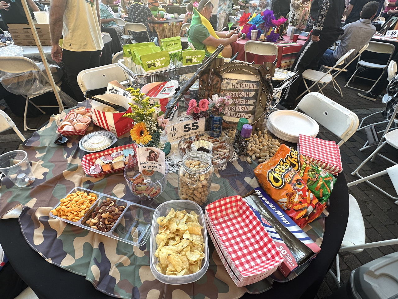 a table with chips, KFC,  peanuts, sauces, etc. There is a camoflauge table cloth underneath all the food with red picnic tablecloth like napkins and plates. 