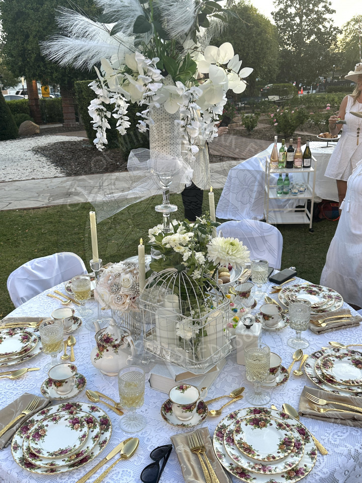 white flowers and greneery with candles surrounding the center of the table. There are floral plates and tea cups around the table and gold silverware.