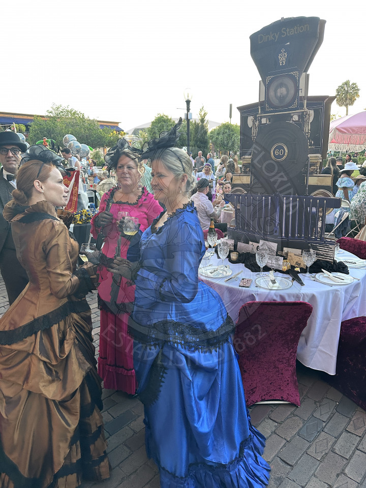 3 women standing in a circle holding a glass with wine in it wearing long dresses from the 19th century and there is bustles. 