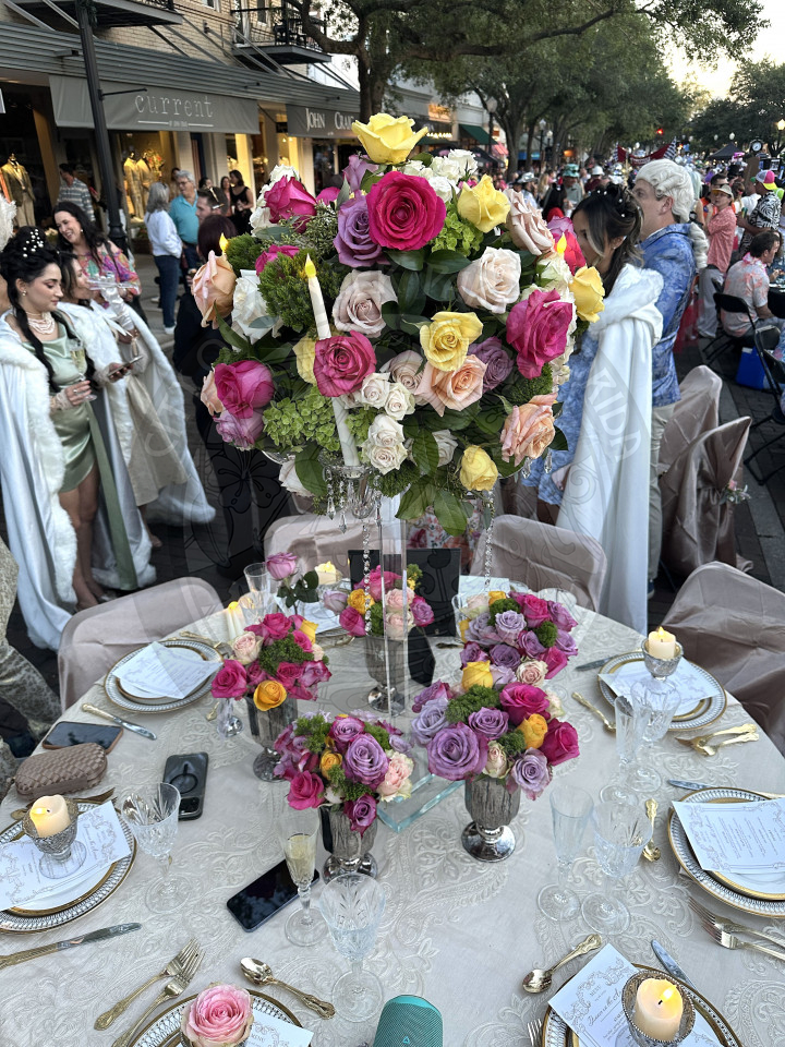 Floral center piece with colorful flowers of hot pink, yellow, purcple, and light pink. they have a neutral floral themed table cloth on the table with table setting of white plates and gold silverware.