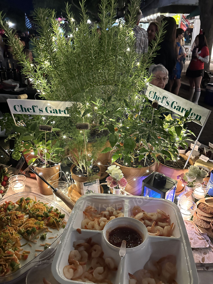 a table with herbs and plants in the middle with signs that say "chef's garden" around the plants are platters of shrimp and sandwiches.