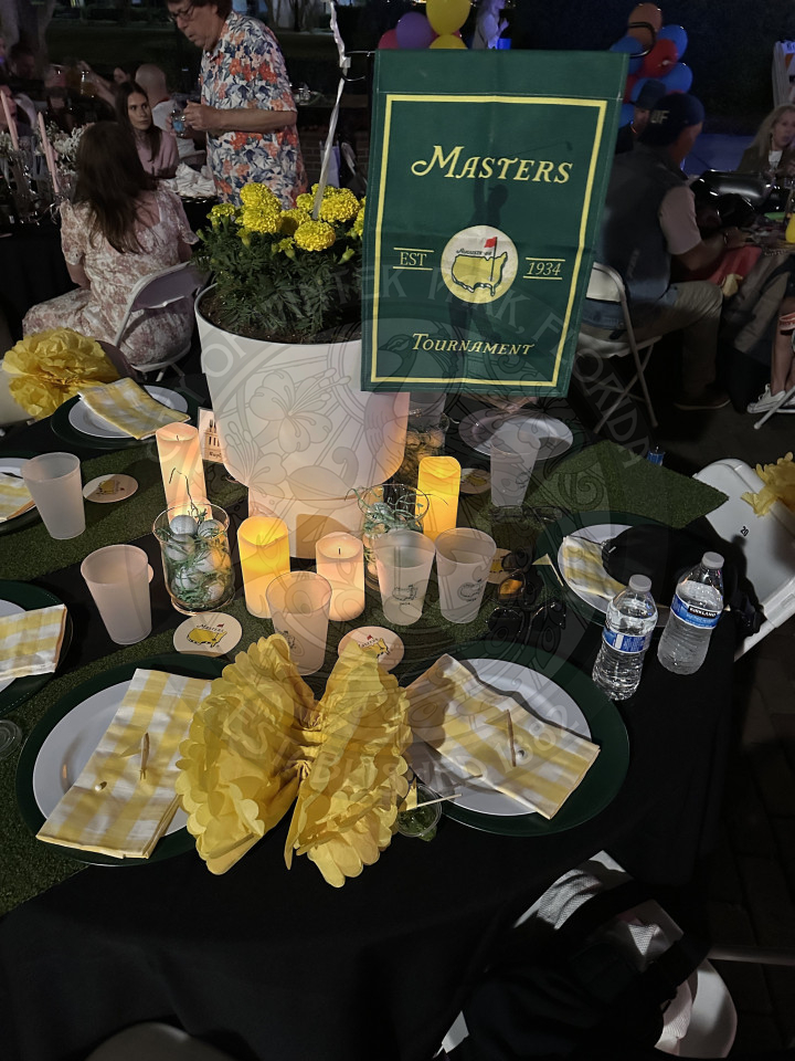 a table with a potted plant in the middle and a garden flag that is logo of the Master's Tournament with  themed costers, cups, and plates on the table.