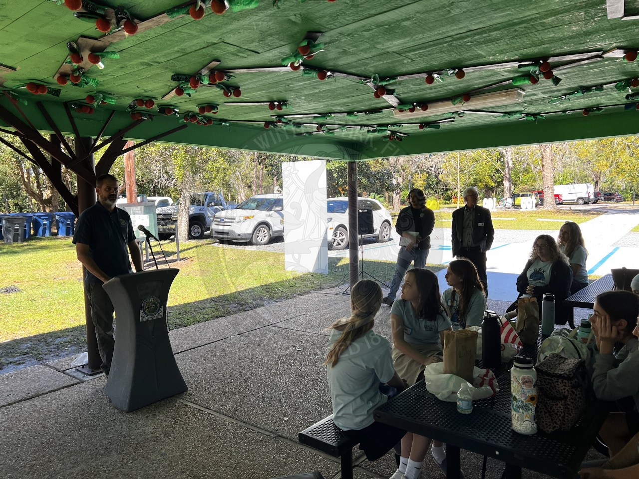 City staff member standing behind podium under pavilion talking to kids sitting on picnic tables listening to him talk.