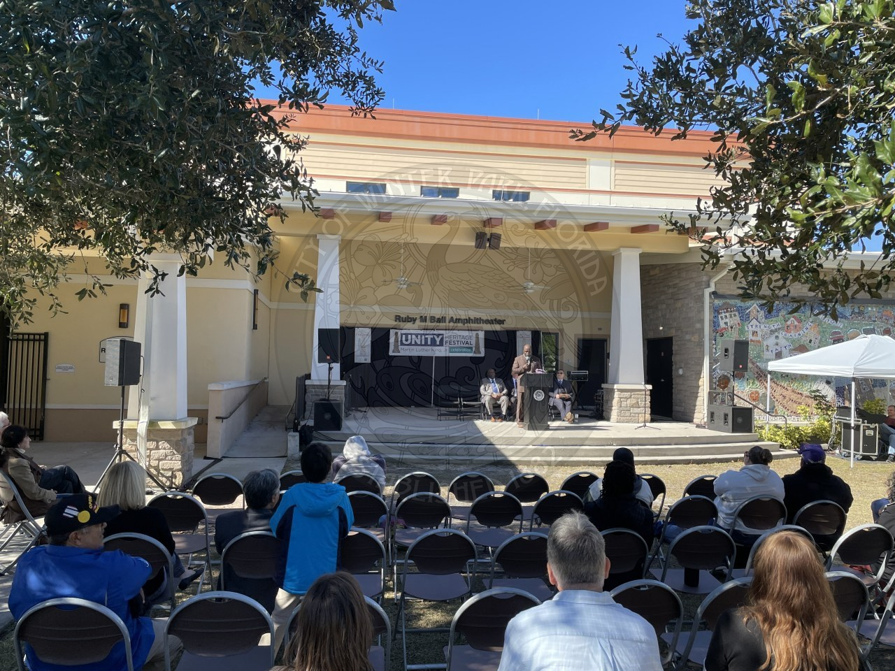 A man standing on stage in front of podium talking to a crowd with people listening to him.