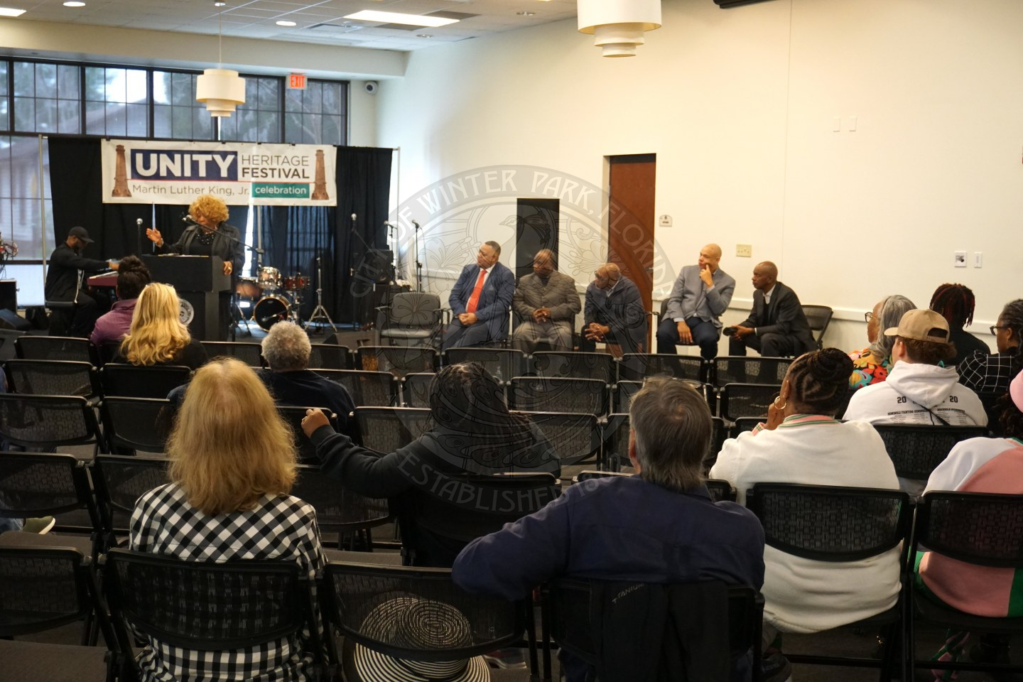 People sitting in rows of black folding chairs with a person in front of evderyone standing behind a podium talking to the audience. 