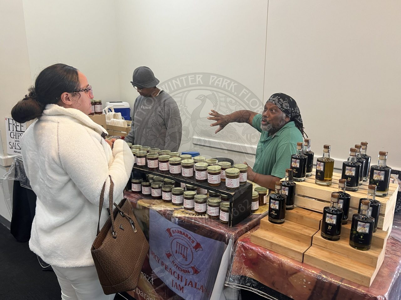 a man sitting behind a table filled with products and talking to someone standing on the other side of the table.