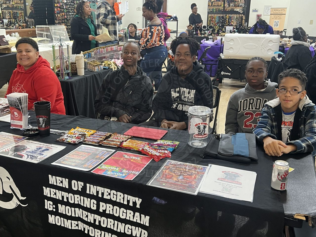 Five men sitting behind a booth with a table cloth that says "Men of Integrity Mentoring program"