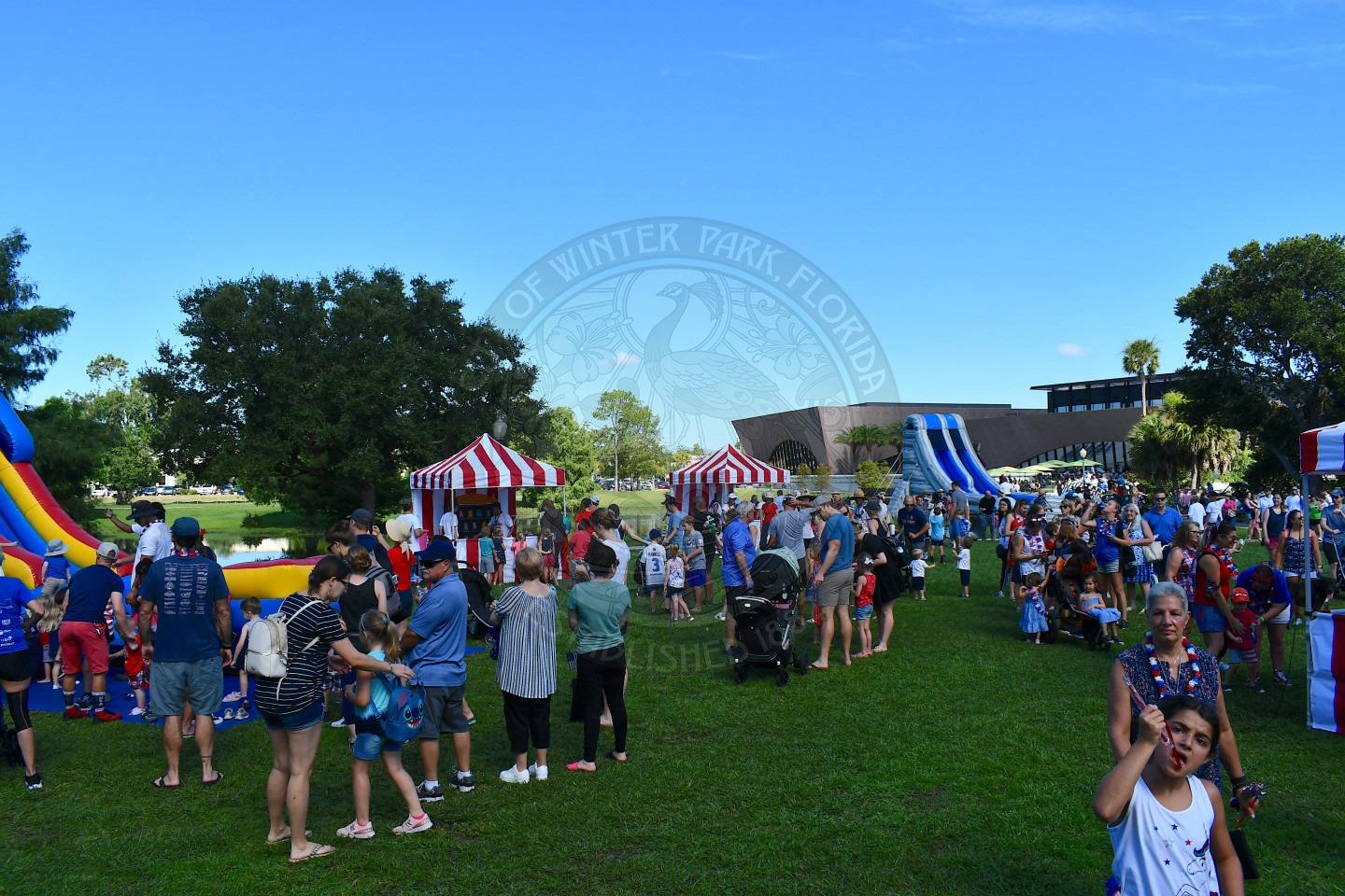 Crowd of people standing around the kid zone which includes carnival tents with games in them and bounce houses.