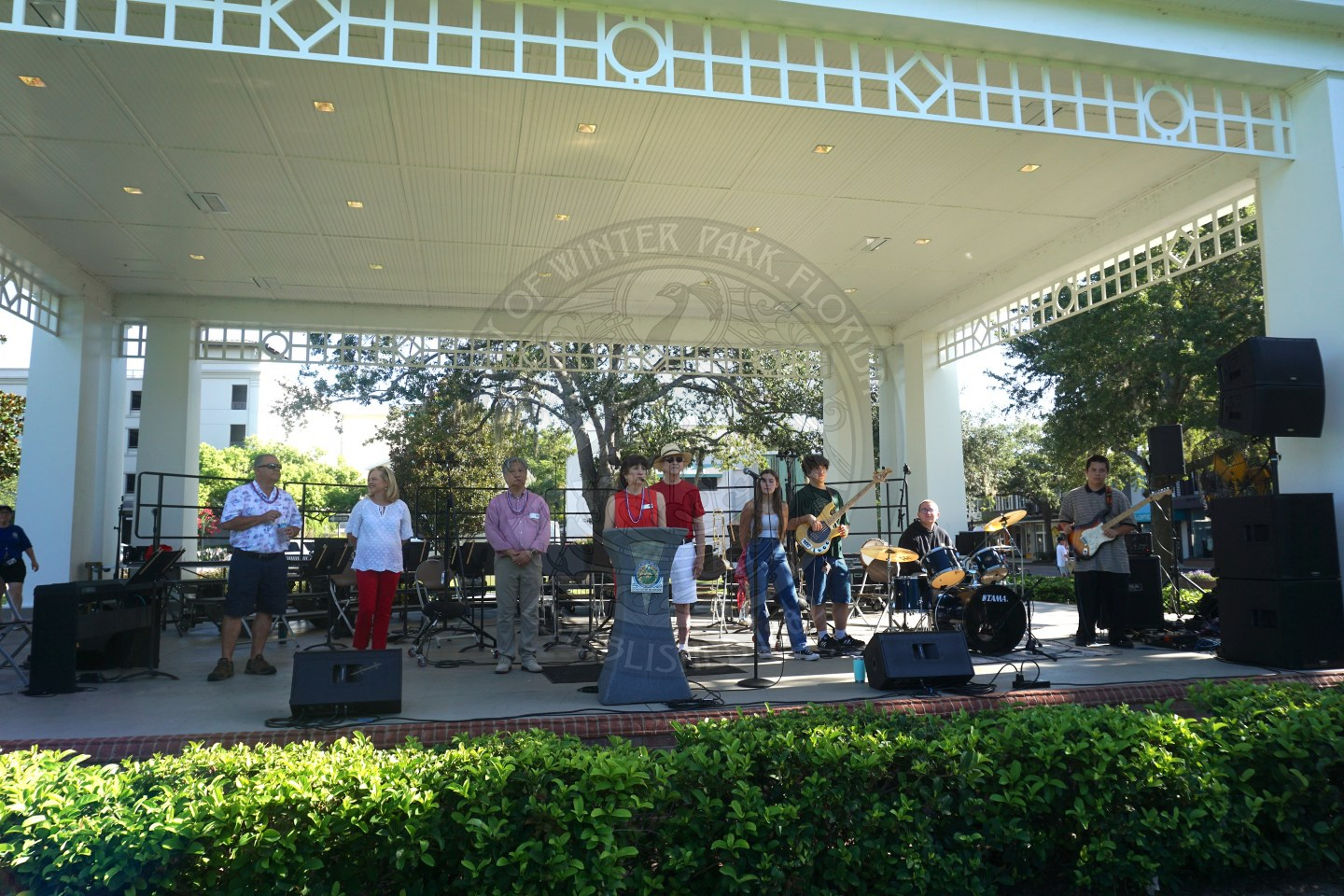 Mayor of Winter Park speaking in front of a podium to the attenedees of the Fourth of July celerbation. Thre is a band with drum set, guitar, and a signer to the right of her.