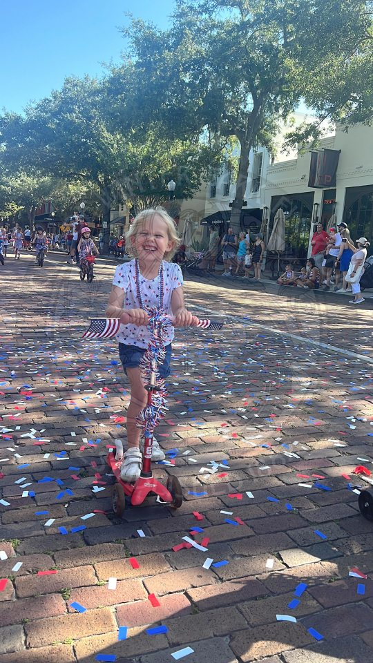a little girl wearing red, white and blue riding her scooter that is decorated with american garland.