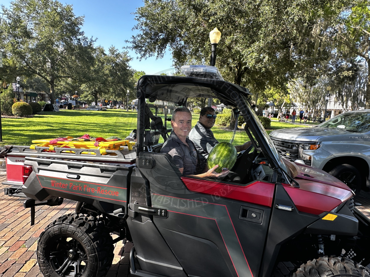 2 firefighters sitting in the golf car while the passenger seat is holding a watermelon.
