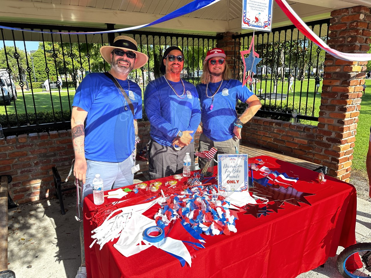 3 men wearing blue city shirts standing behind a table with fourth of july decorations on it.