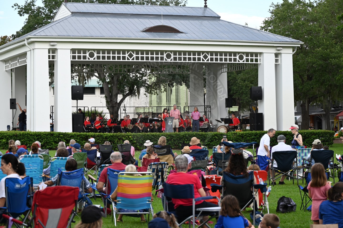People sit on lawn chairs watching a band perform on an outdoor stage in a park with trees and a pavilion.