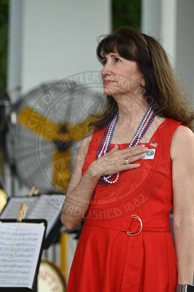 Woman in red dress stands with hand on chest, wearing patriotic beads, during an outdoor event, music sheets visible.