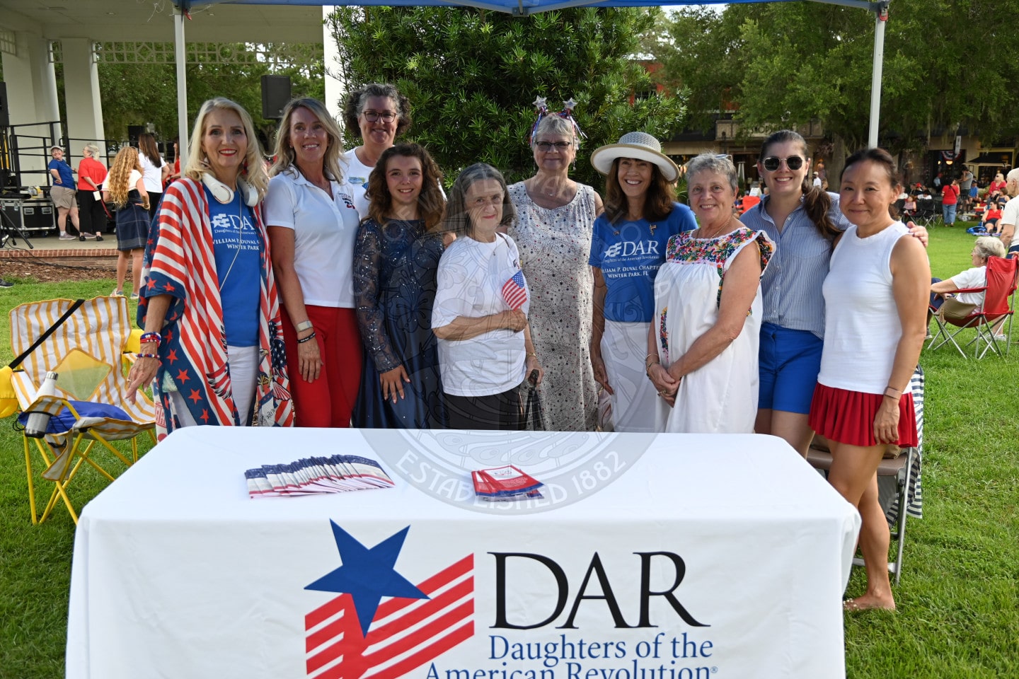 Ten women stand behind a DAR table outdoors, smiling; some wear patriotic colors and hold small American flags.