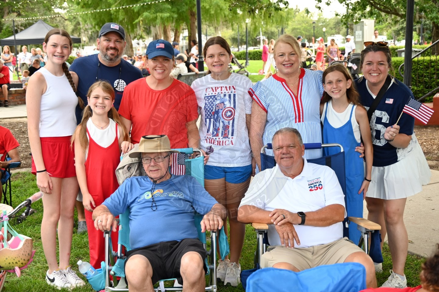 A group of people in patriotic outfits smile outdoors at a summer event, two men are seated in front.