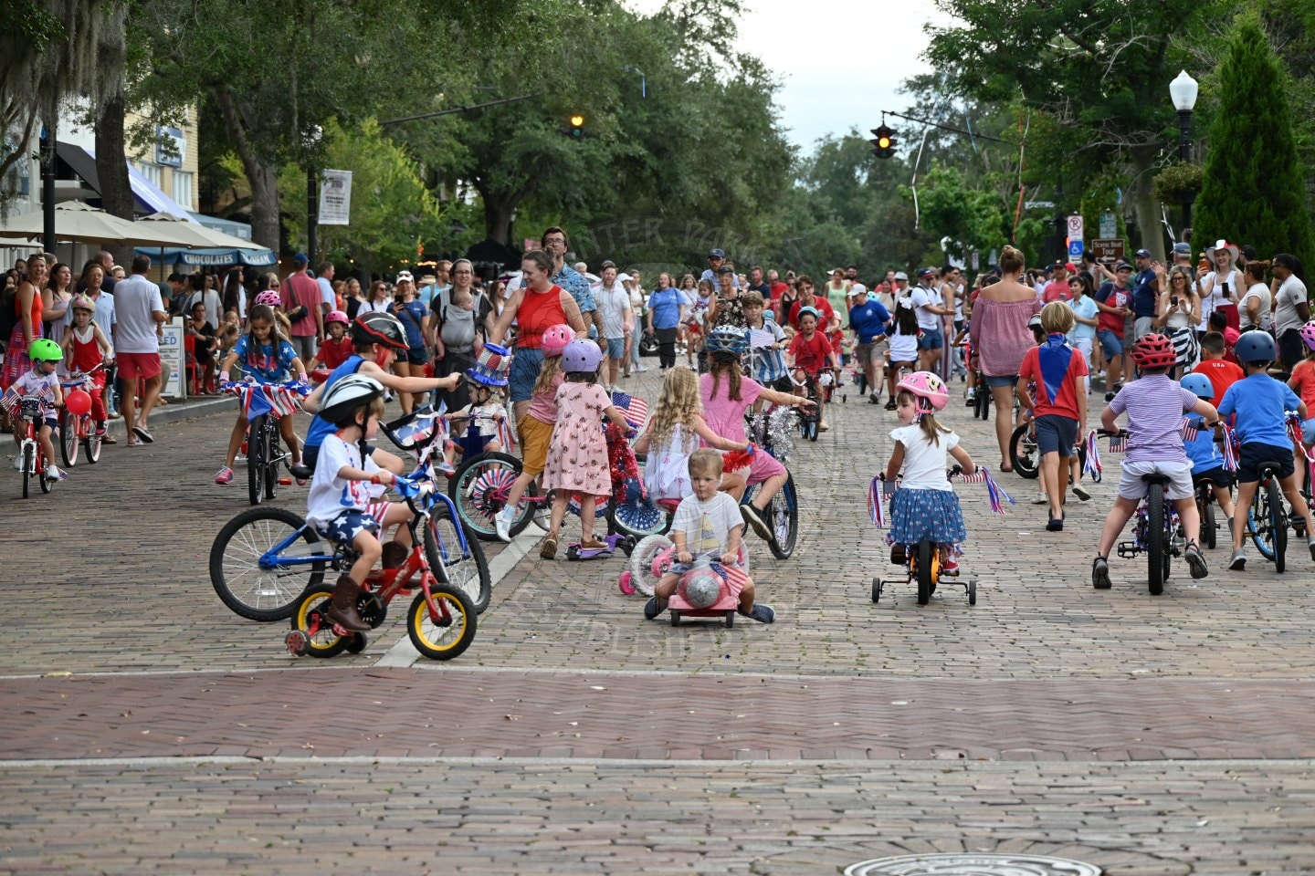Children ride decorated bikes on a crowded street, surrounded by people on foot, during a festive event.