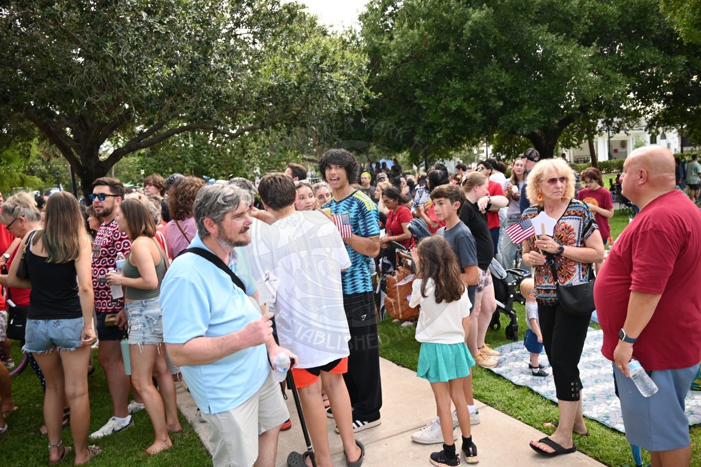 People of all ages gather outdoors on grass, some holding flags, enjoying a community event under trees.