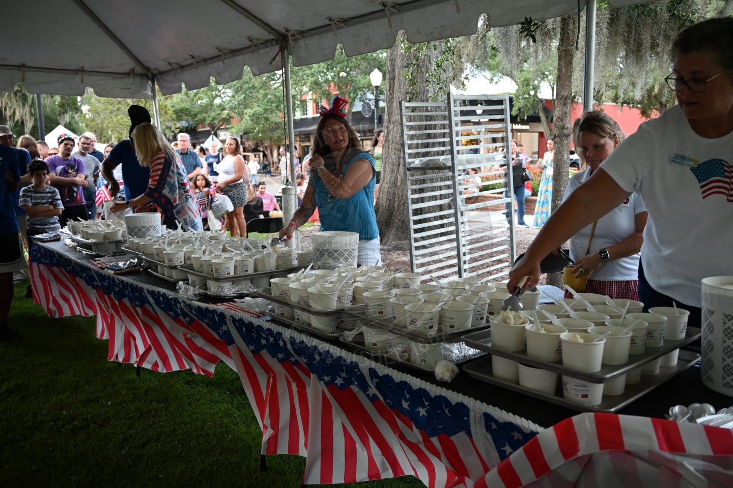 People serve cups of ice cream on tables decorated with American flags at an outdoor event under a tent.