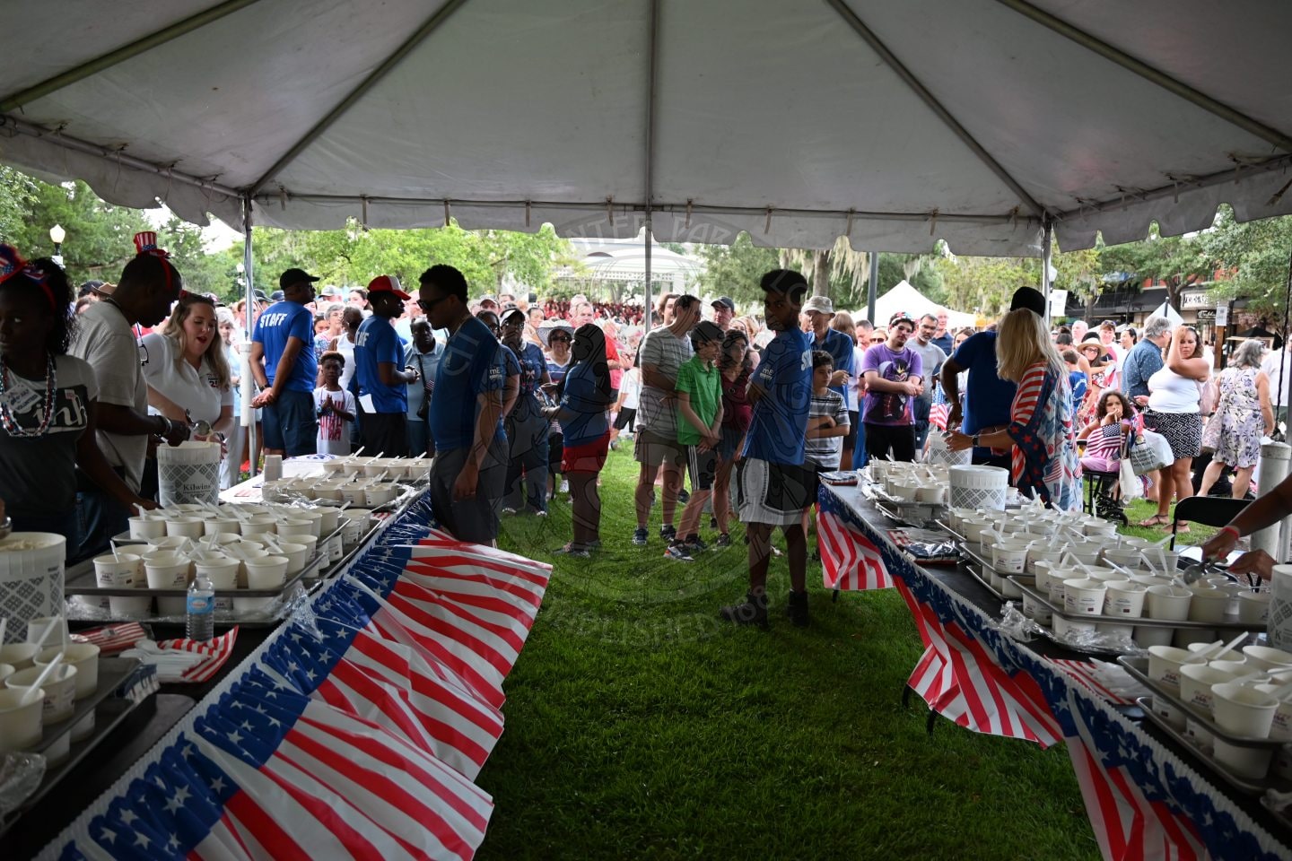 People line up under a tent with tables of ice cream and cake, decorated with American flag tablecloths, at an outdoor event.